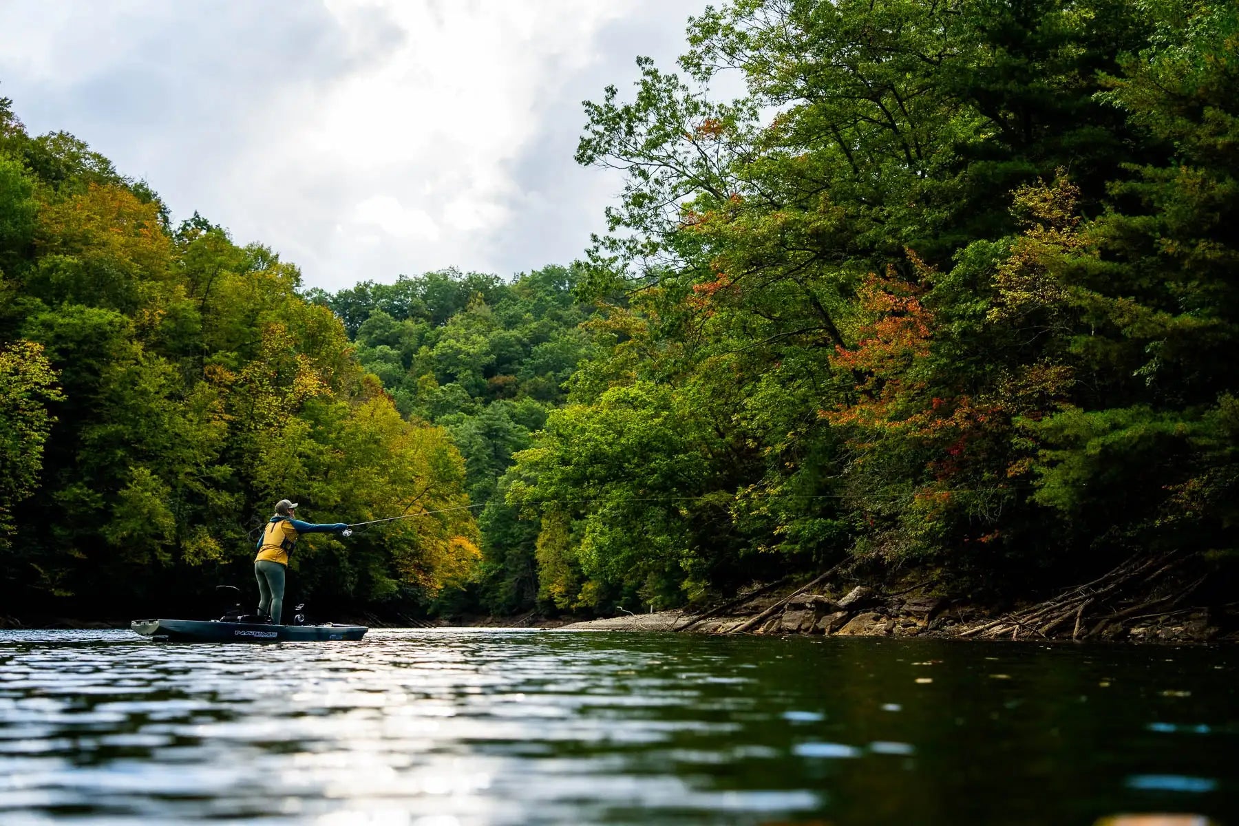 A person in a yellow life vest stands on a dark blue inflatable kayak, casting a fishing line into the water.